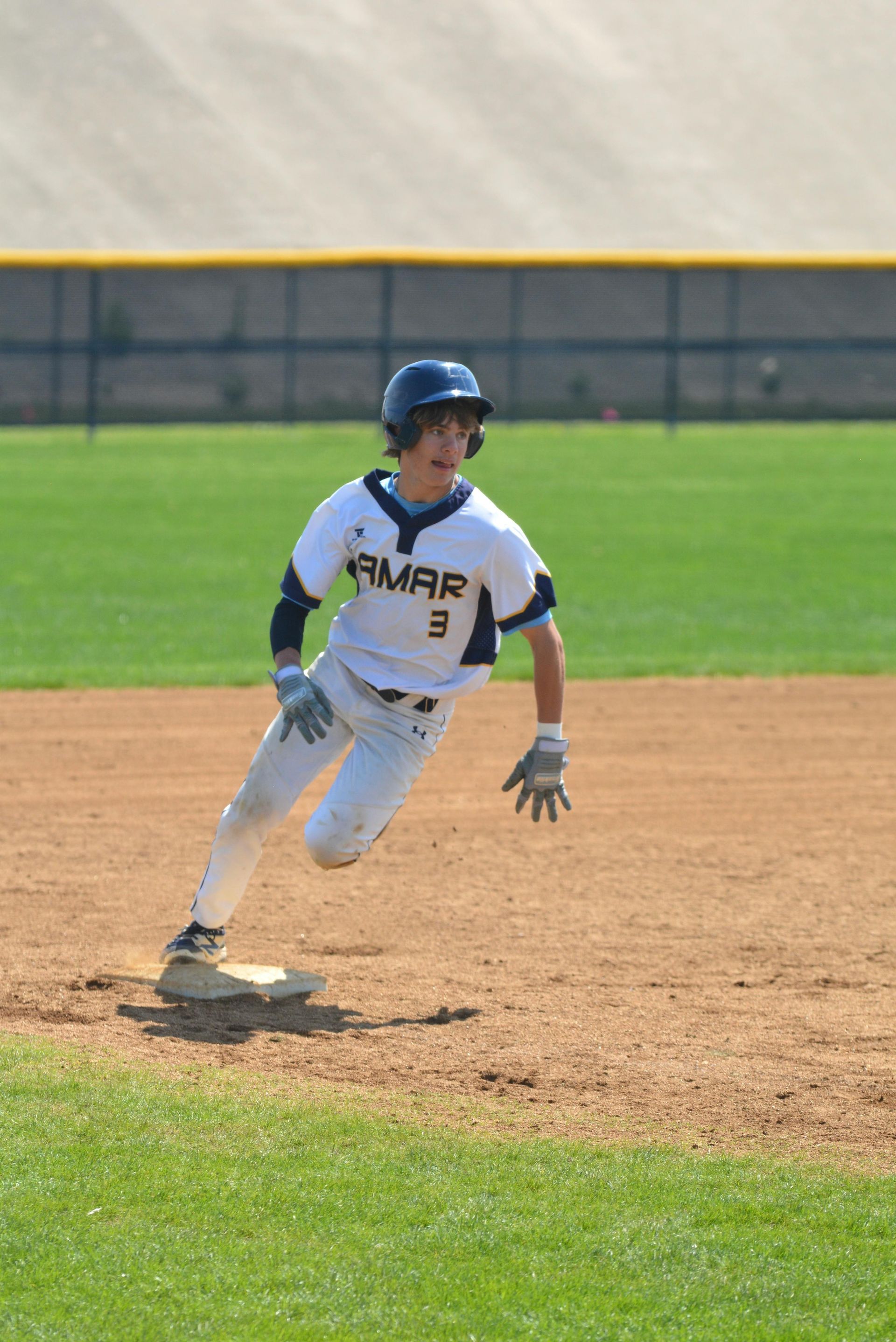 A baseball player in a white and blue uniform leads off a base on a dirt diamond under a sunny sky.