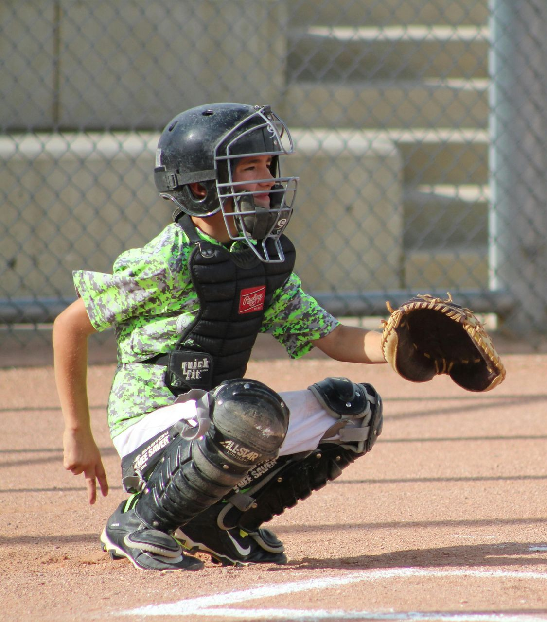 A catcher wearing green patterned gear and protective equipment crouches on a baseball field, signaling with their hand.