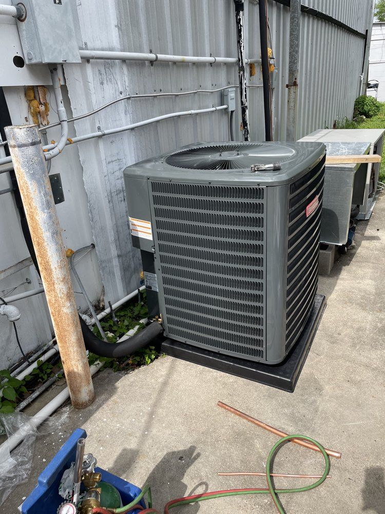 A gray Goodman central air conditioning unit sits on a concrete pad next to a corrugated metal building.