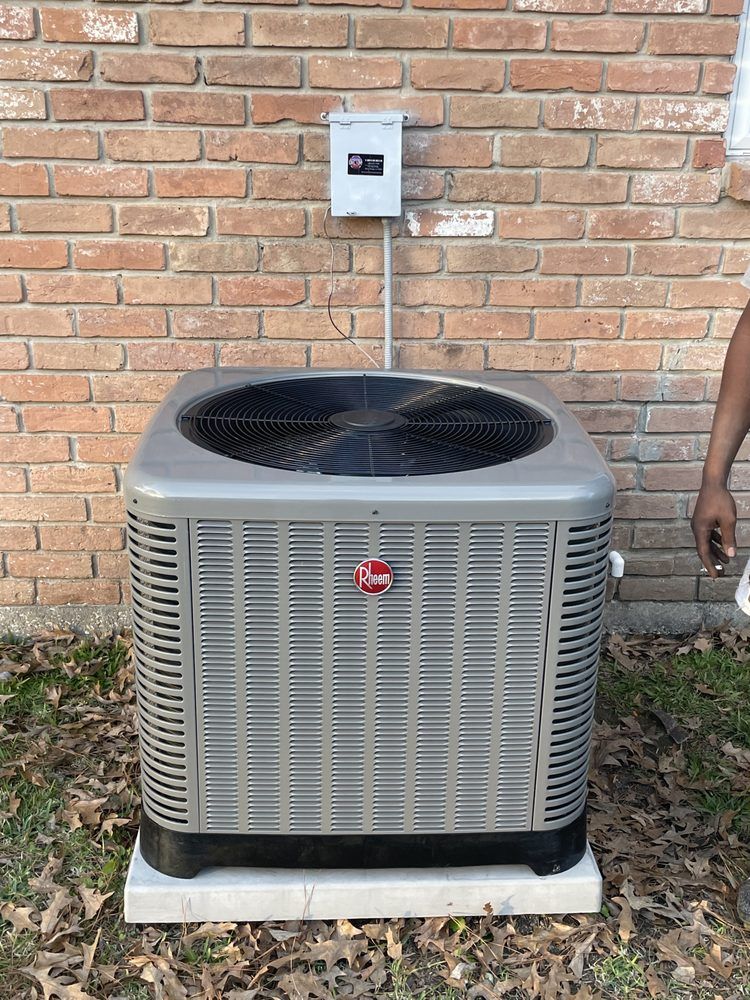 A grey Rheem outdoor air conditioning unit sits on a white concrete pad against a brick wall with an electrical box above.