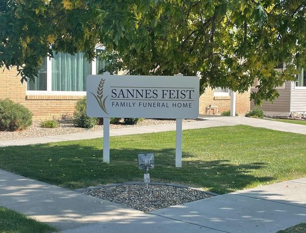 Sannes Feist Family Funeral Home sign on a lawn beside a sidewalk, shaded by trees
