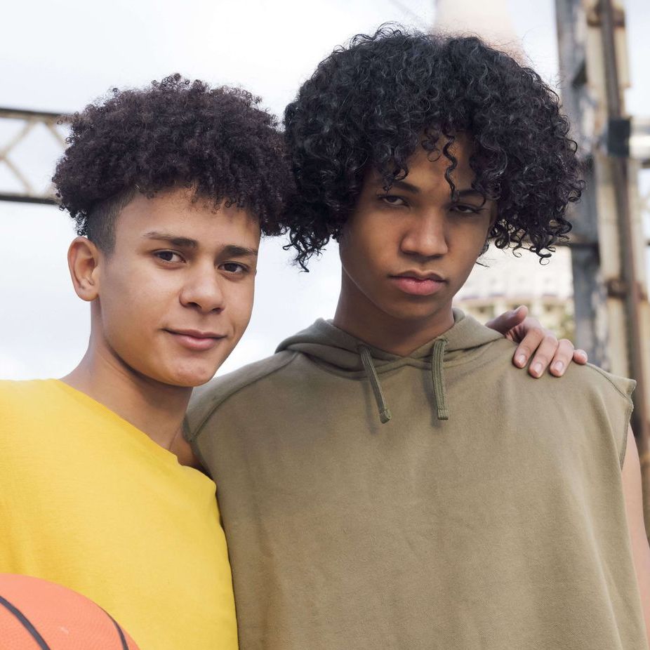 Two young men standing outdoors, one in yellow smiling, other in green hoodie, arm around the other.