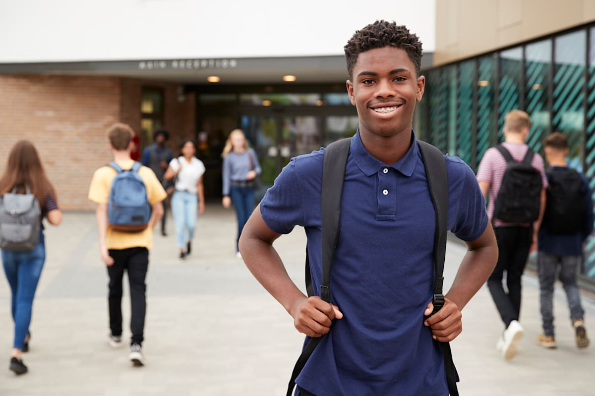 Teenage boy with backpack smiles at the camera outside a school entrance.