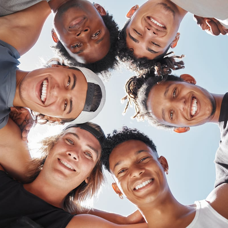 Group of smiling diverse young men, looking down at the camera in a circle, against a blue sky.