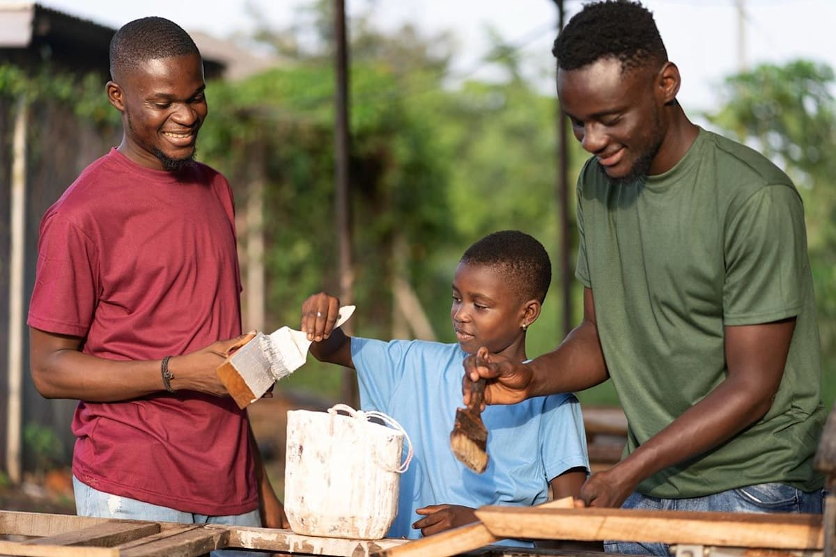 Three Black males, two adults and a child, working together outdoors, smiling, and holding materials.