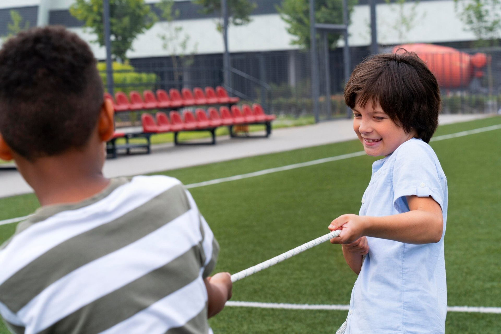 Two boys playing tug-of-war on a green field. The boy in the foreground smiles, pulling a white rope.