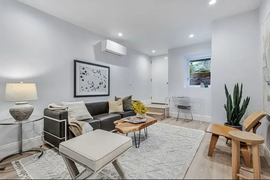 Bright modern living room with gray sofa, white rug, wooden tables, wall art, and potted plant