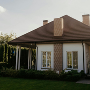 Single-story house with brown roof, brick chimney, white walls, and front windows amid green landscaping