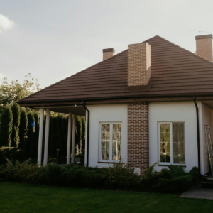 Single-story house with brown roof, brick chimney, white walls, and front windows amid green landscaping