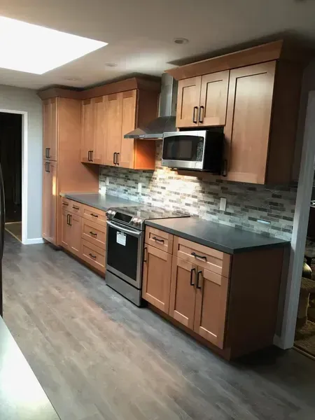 Empty kitchen with wooden cabinets, gray backsplash, black appliances, and wood-look flooring.