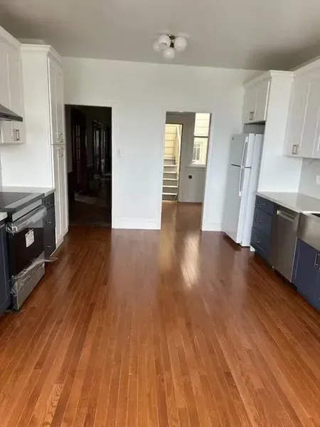 Empty white kitchen with wood floors, black appliances, and a refrigerator near a doorway