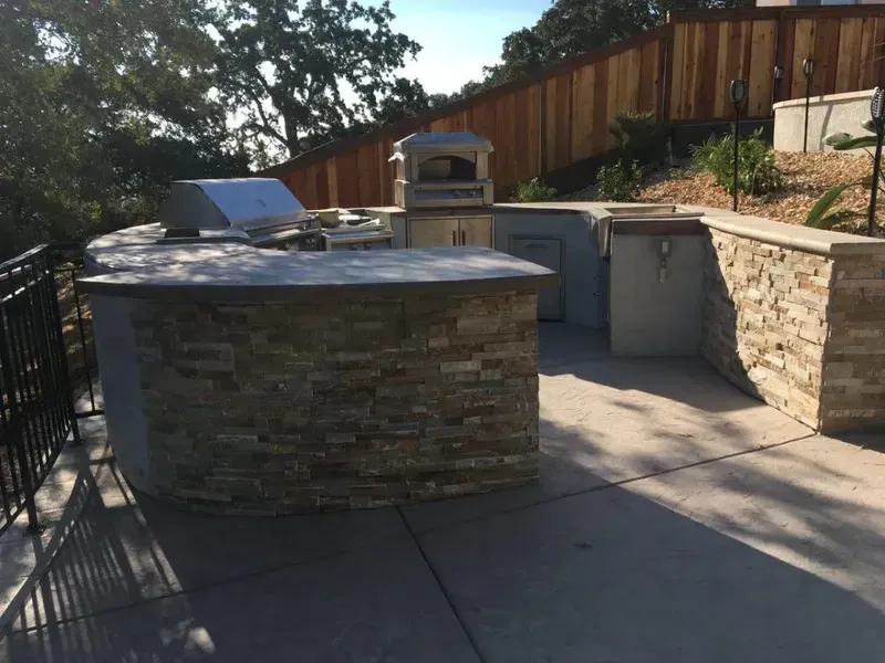 Outdoor stone patio with built-in grill and countertops beside a sloped wooden fence