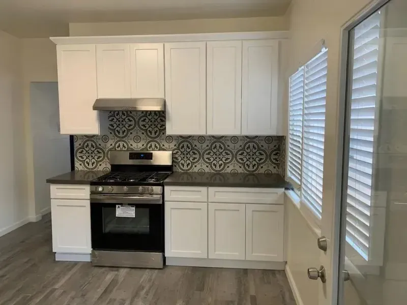 Small modern kitchen with white cabinets, black stove, patterned tile backsplash, and windows with blinds