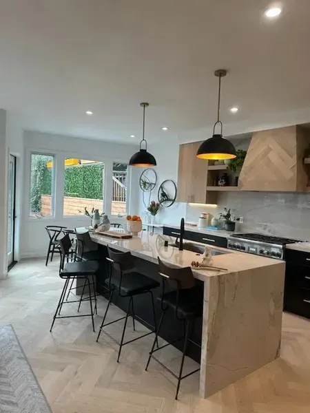 Modern kitchen with island, black bar stools, pendant lights, and large windows overlooking greenery