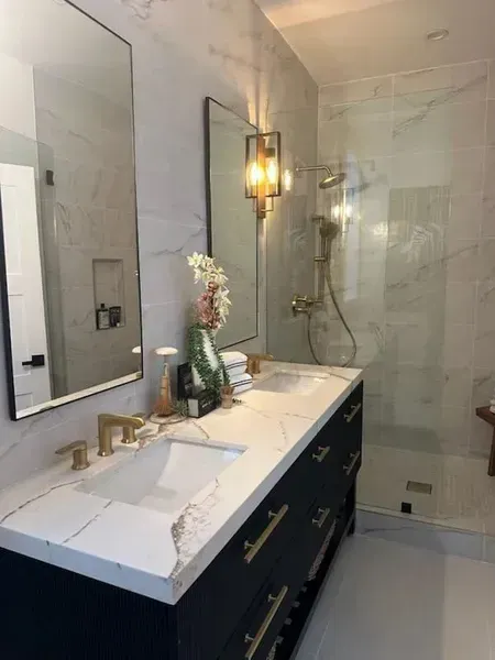 Modern bathroom with double vanity, brass fixtures, large mirror, glass shower, and dark blue cabinets