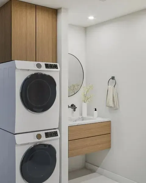Modern laundry bathroom with stacked washer-dryer, wood vanity, round mirror, and white towel on wall.