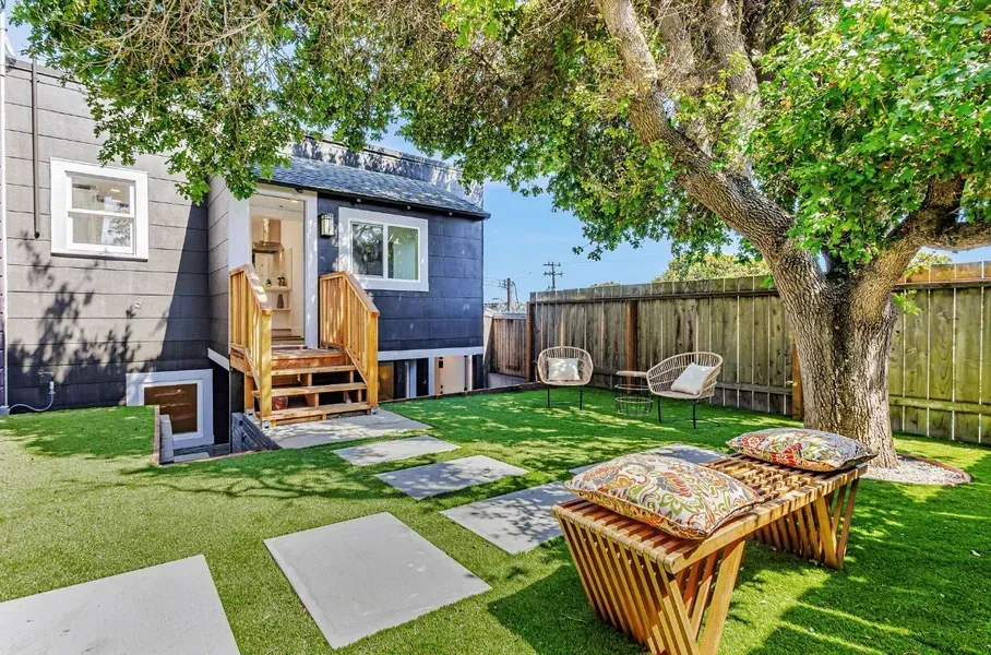 Backyard patio with green lawn, wooden deck, wicker chairs, and a tree beside a small blue house