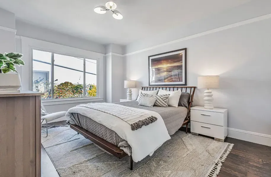Bright modern bedroom with a bed, nightstands, lamps, large window, and soft gray decor