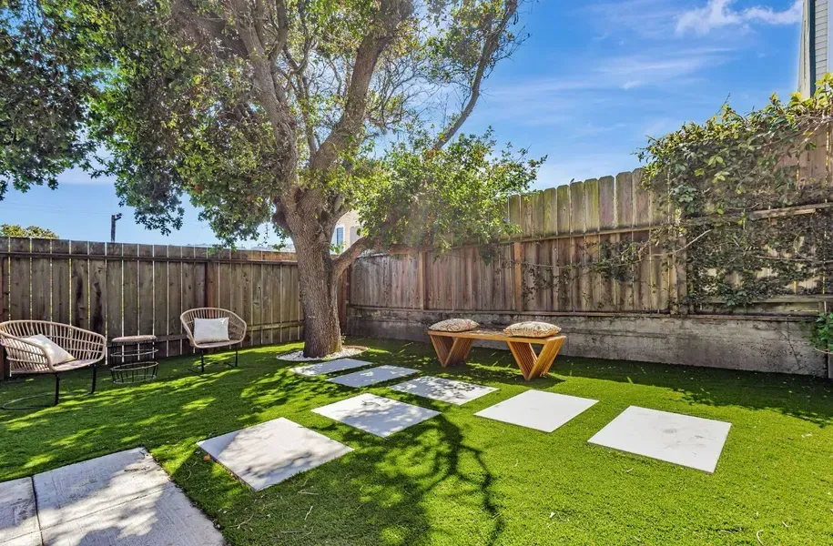 Sunny backyard with green lawn, stepping stones, a large tree, and two wooden chairs by a fence