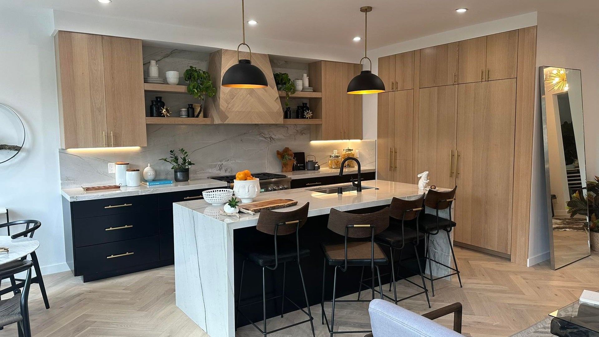 Modern kitchen with light wood cabinets, black island stools, pendant lights, and a dining table by a window.