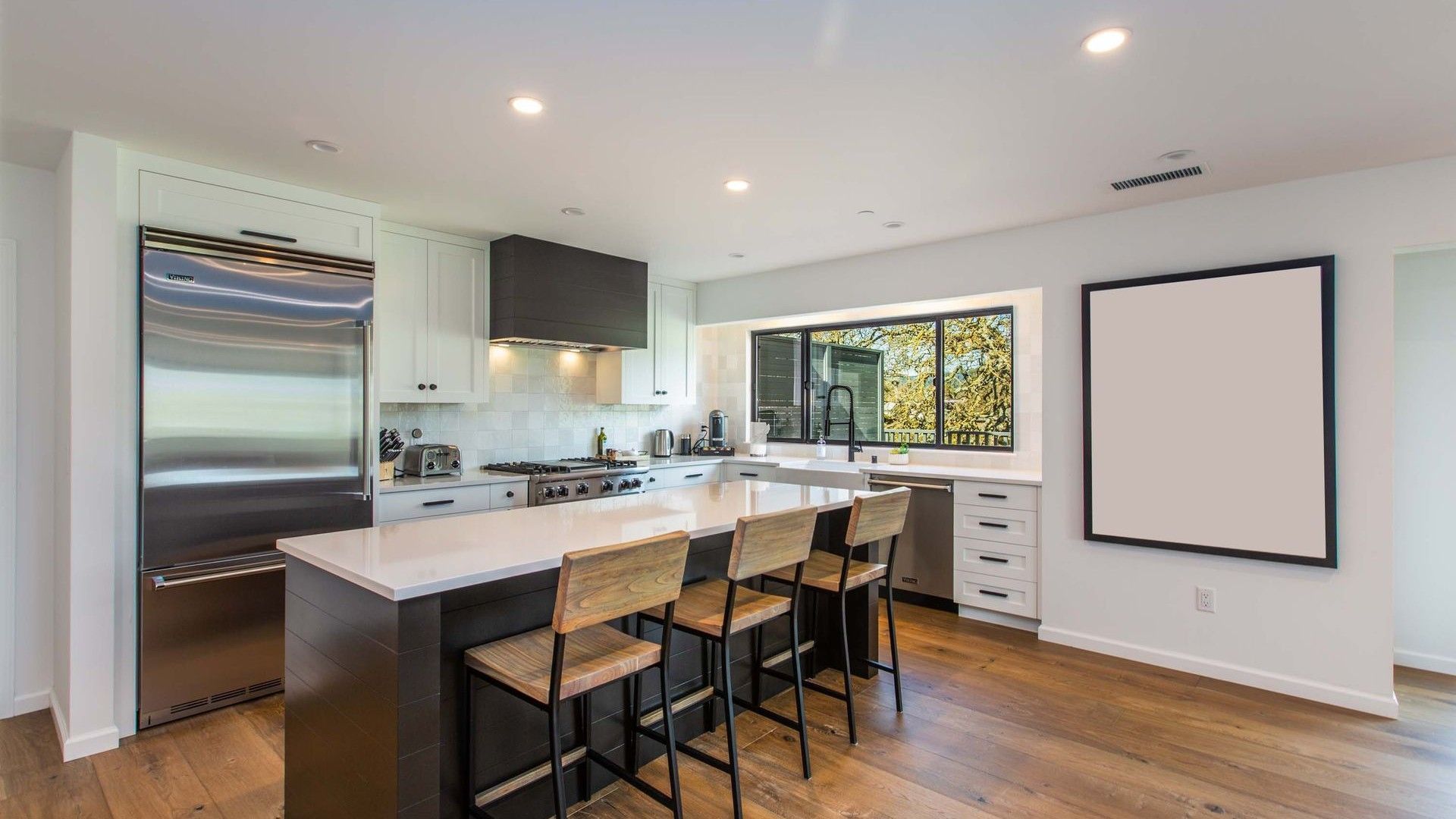 Modern kitchen with island, bar stools, stainless appliances, wood floors, and a blank wall-mounted screen