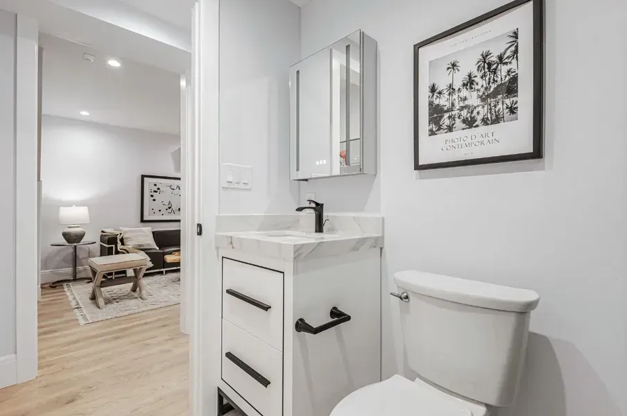 Bright white bathroom with vanity, sink, toilet, and framed art; doorway opens to a cozy bedroom beyond