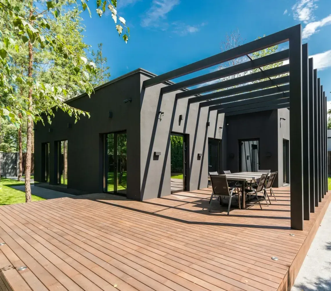 Modern black house with a wooden deck, patio furniture, and pergola in a sunny yard