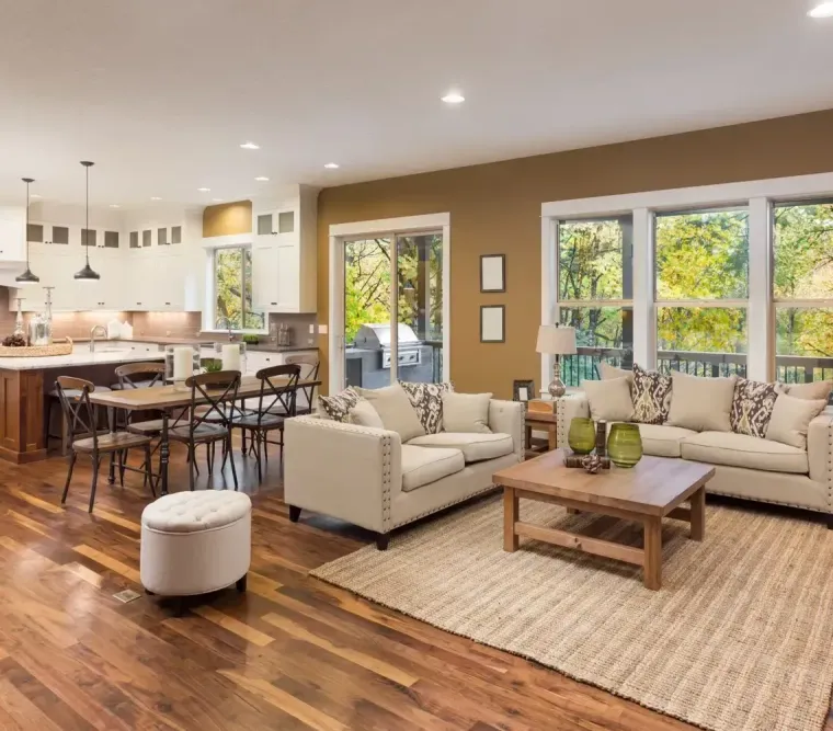 Bright open-concept living room with kitchen, beige sofas, wood floors, and large windows overlooking greenery