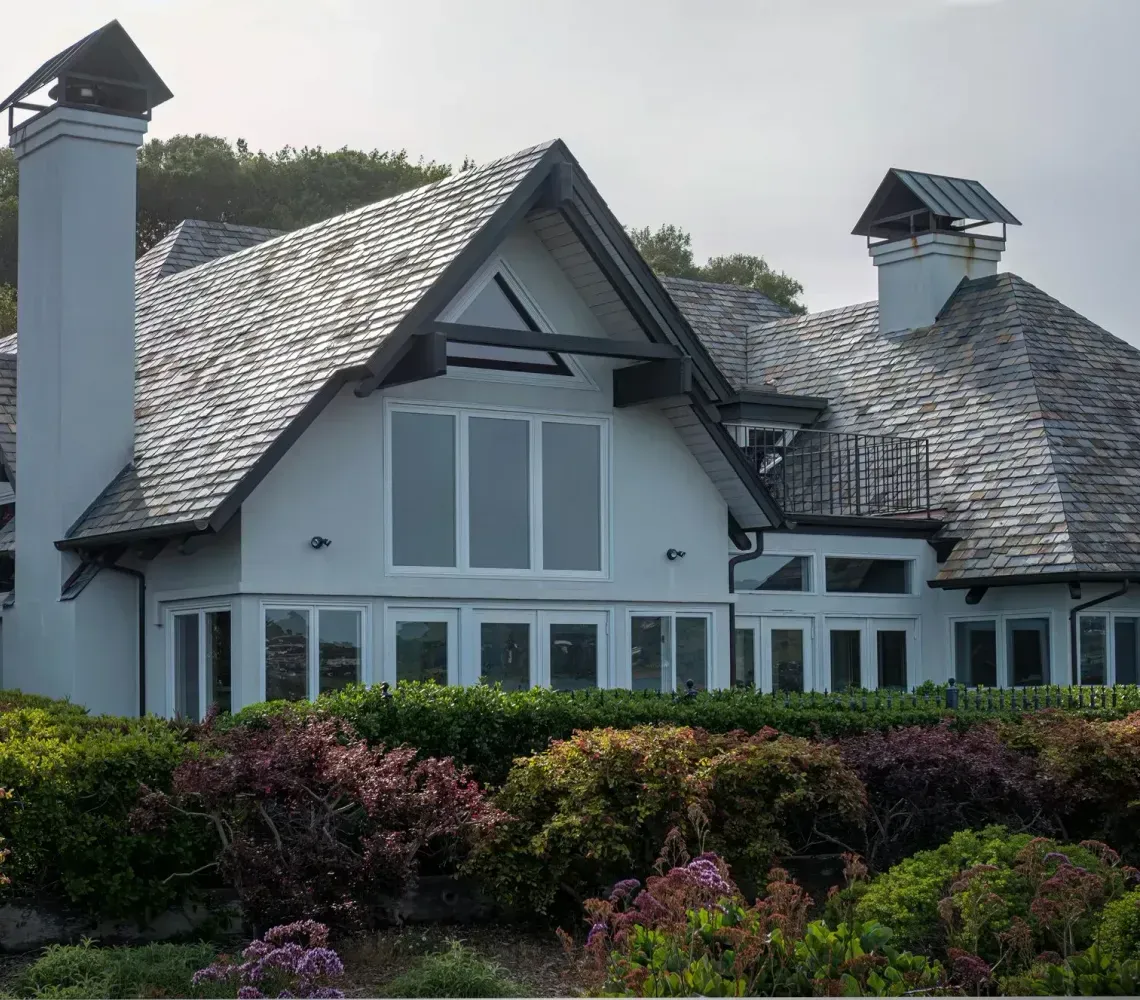 Large white house with gray roof and chimneys, set behind colorful shrubs and trees