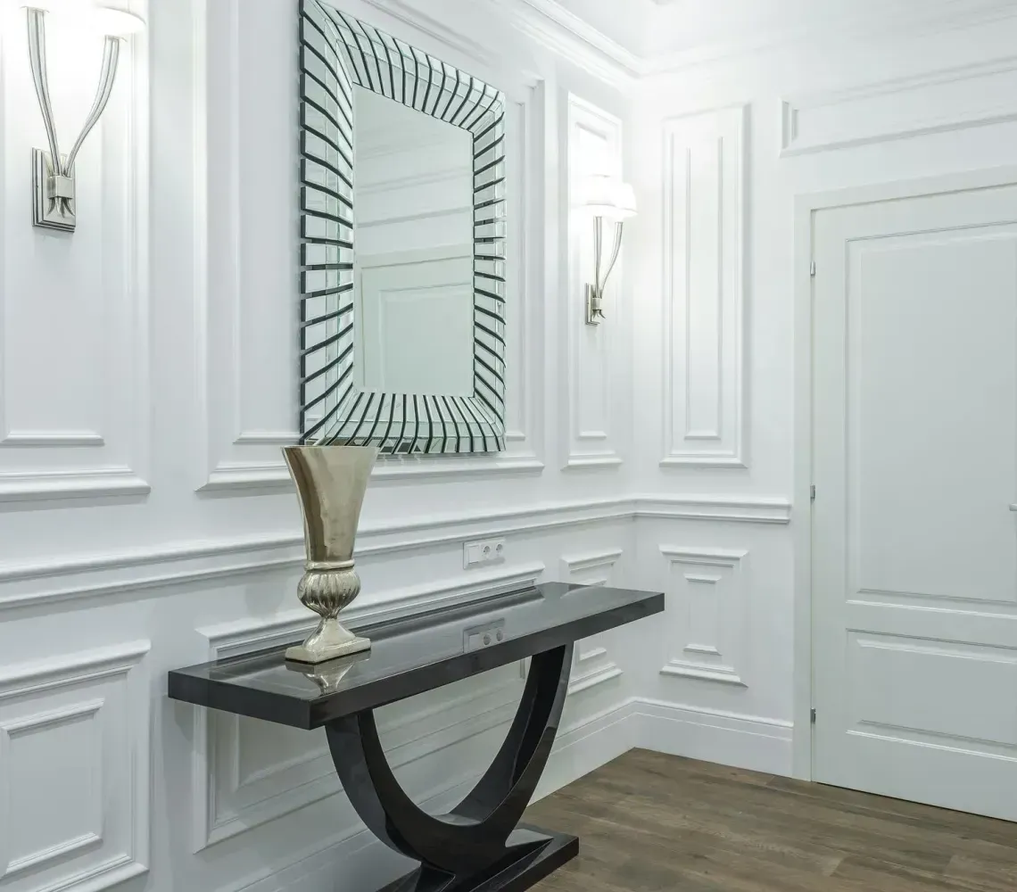 Bright white hallway with paneled walls, a console table, mirror, and closed door