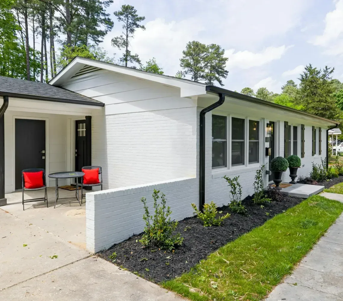 White single-story house with a small covered porch, red chairs, and a concrete driveway with landscaping