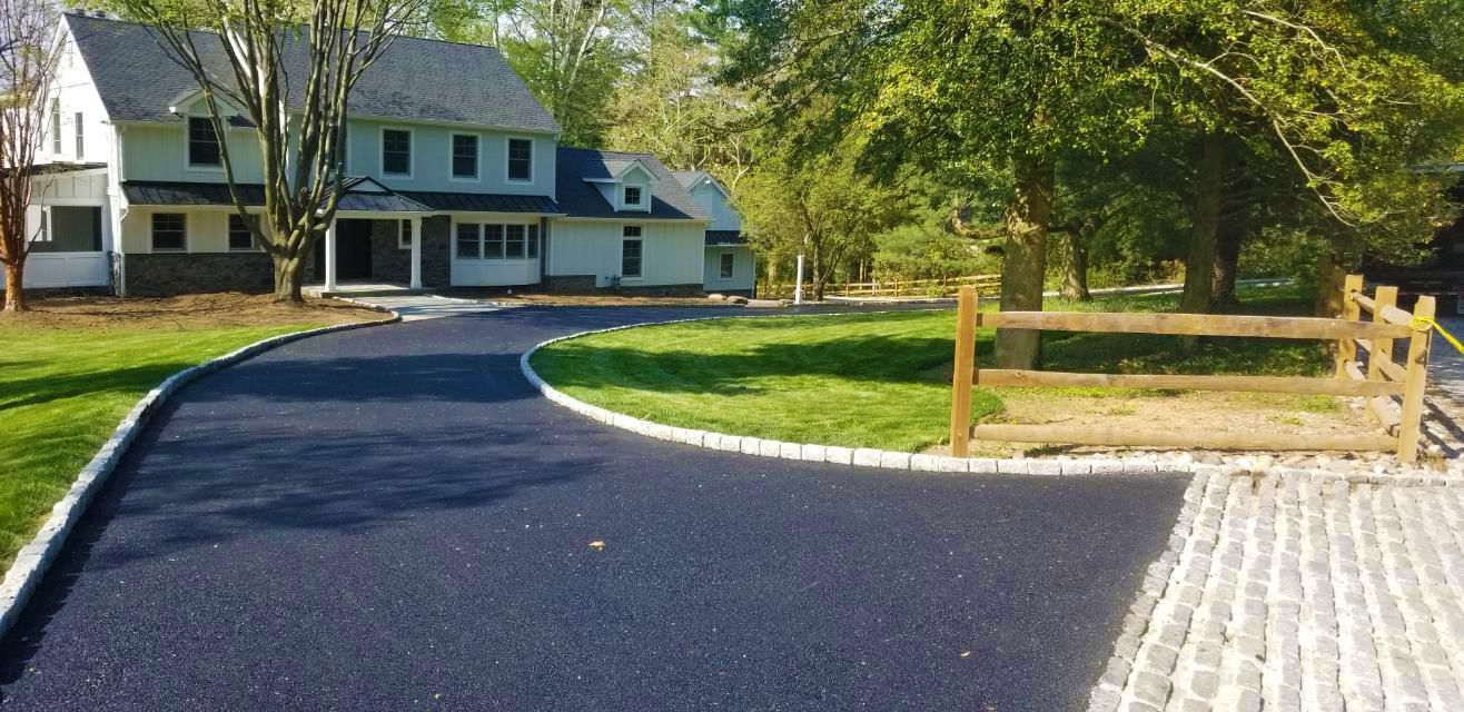 A paved driveway leads to a large white house with a green lawn and wooden fence.