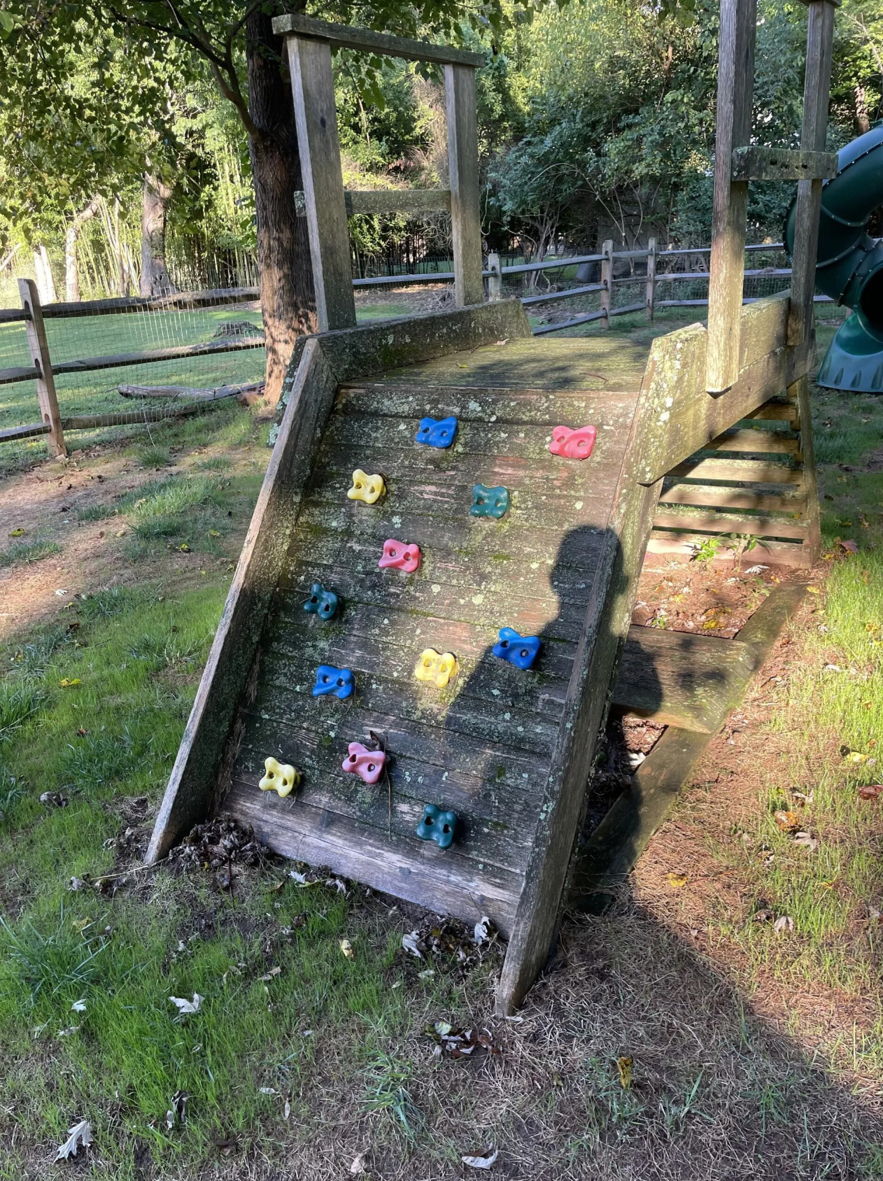Wooden climbing wall on a playground, with colored handholds. Outdoors, with a fence and trees in background.
