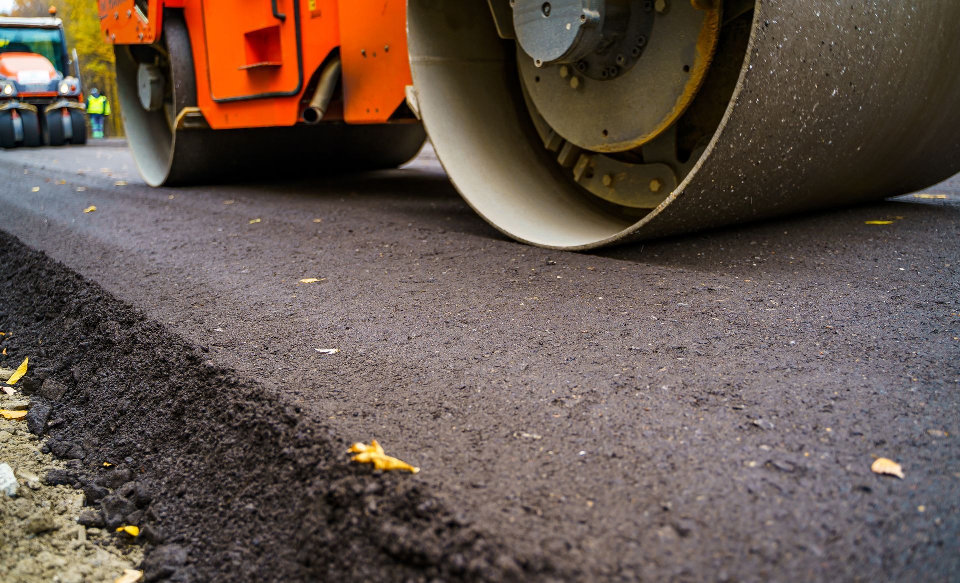 A bright orange industrial road roller compacts a freshly laid black asphalt surface.