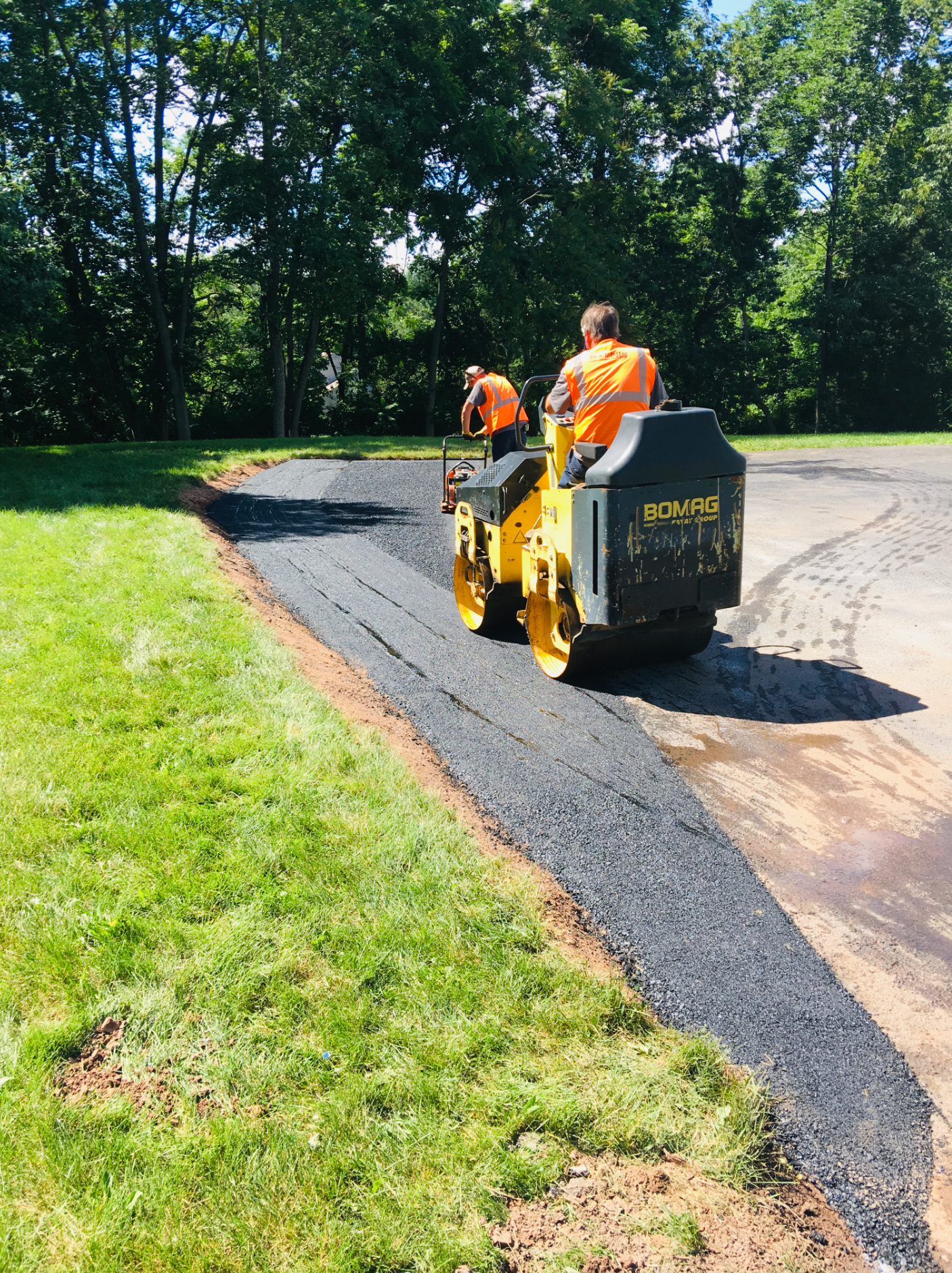 Two workers in orange safety vests operate a yellow asphalt roller to pave a driveway edge next to a grassy lawn.