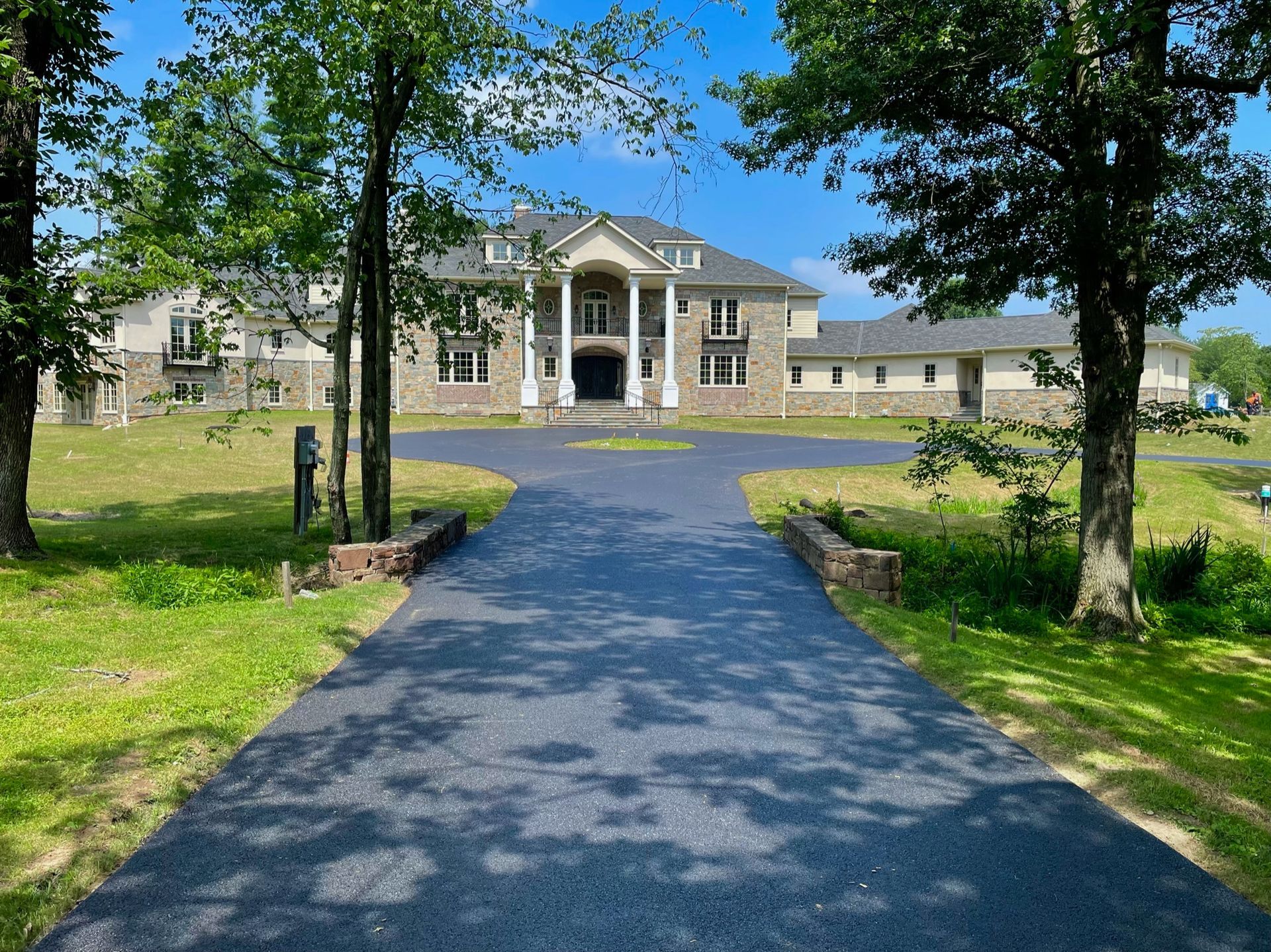 A large stone mansion with a gray roof at the end of a paved driveway, surrounded by green lawns and trees.
