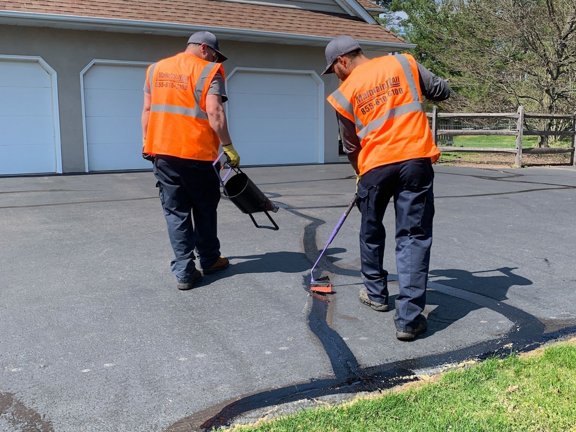 Two workers in orange vests seal a driveway, one using a leaf blower, the other using a brush.