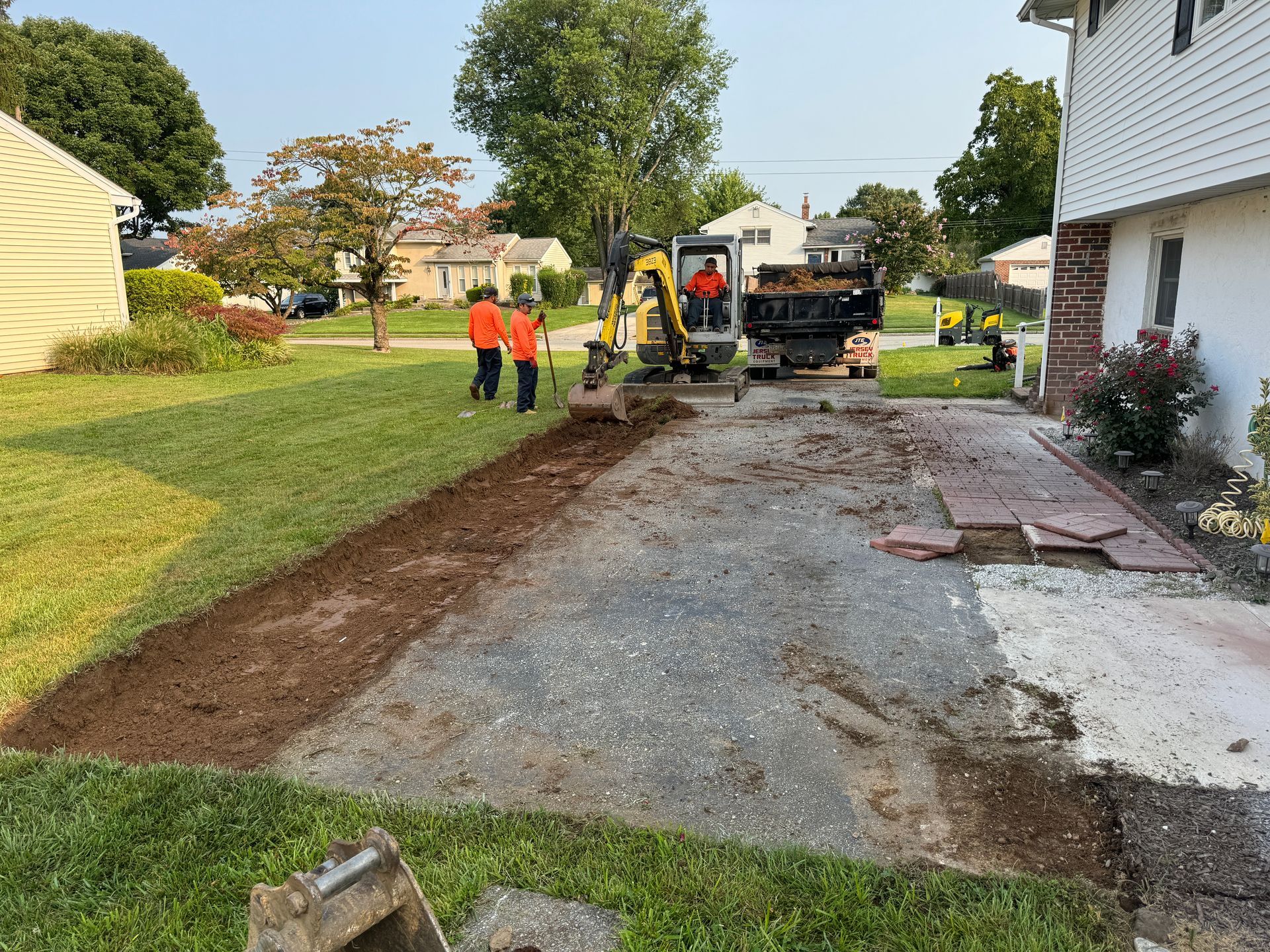 Workers in orange safety gear operate a small excavator on a suburban driveway under construction next to a house.