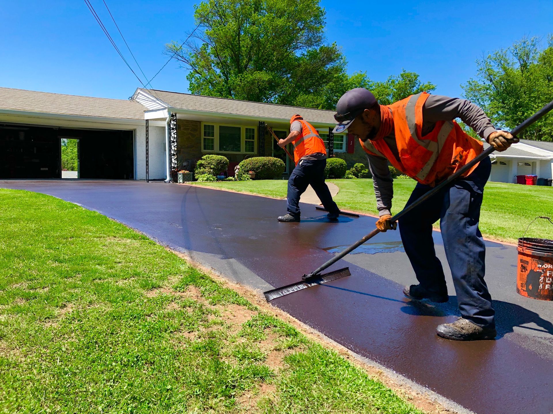 Two workers in orange safety vests use squeegees to apply dark sealant to a residential driveway.