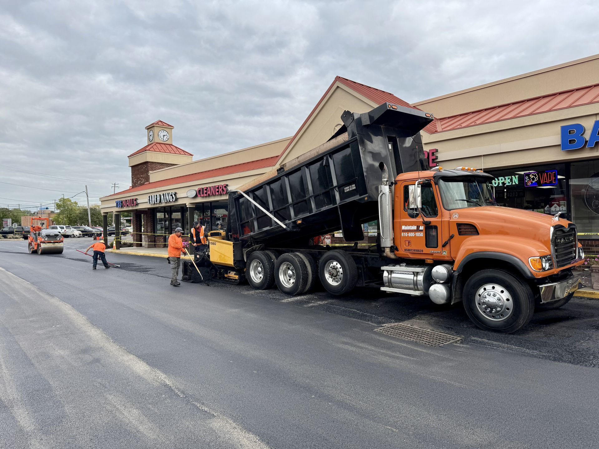 Construction crew paving an asphalt road with a dump truck and a steamroller in an outdoor industrial area.