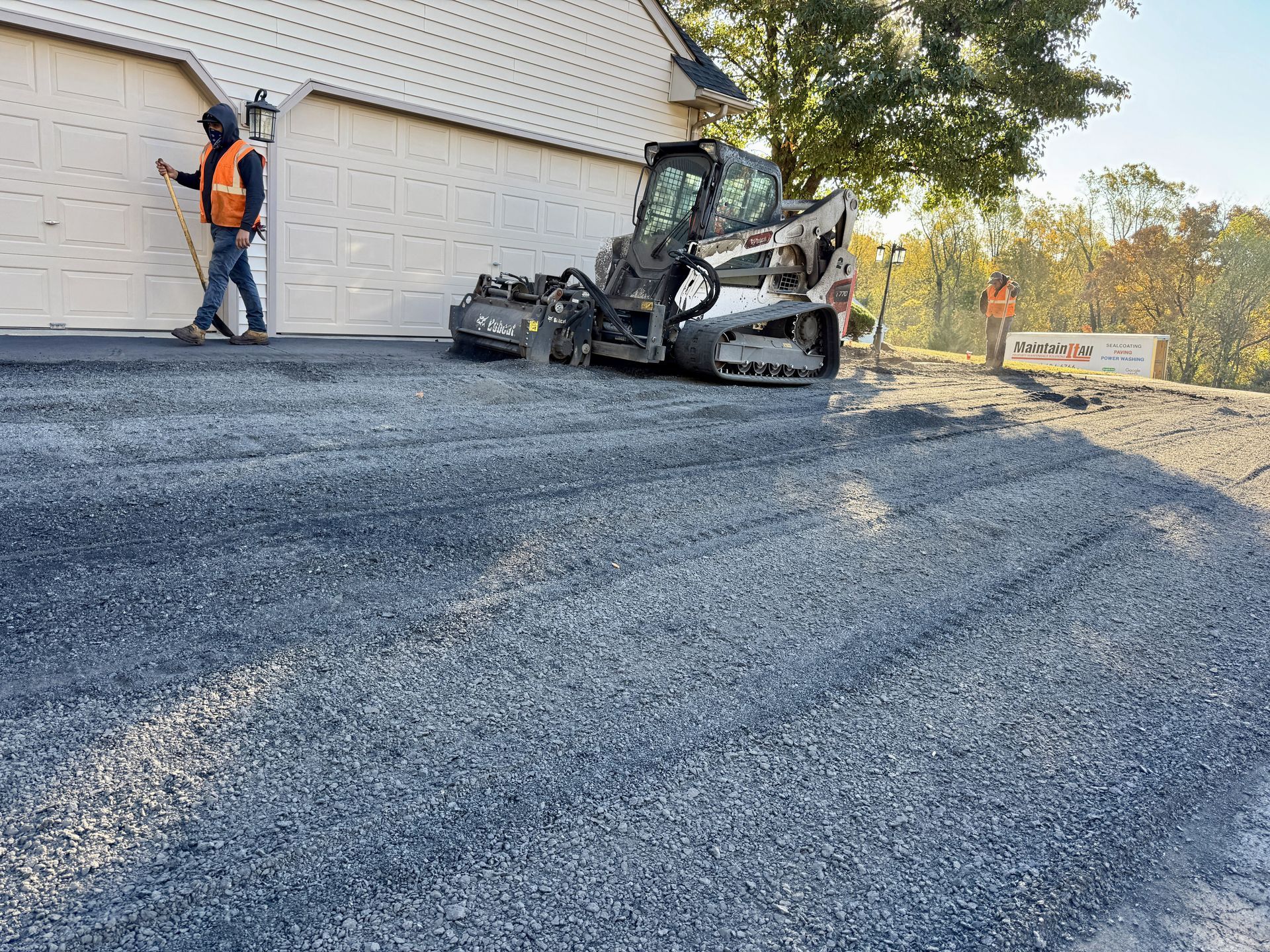 Workers prepare a gravel driveway for paving in front of a residential garage with a skid steer loader nearby.