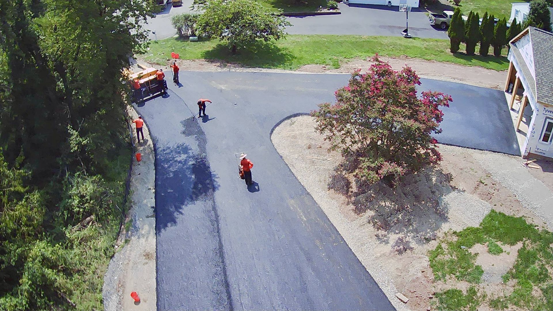 A crew of workers in orange shirts paves a road with asphalt using heavy machinery on a sunny day.