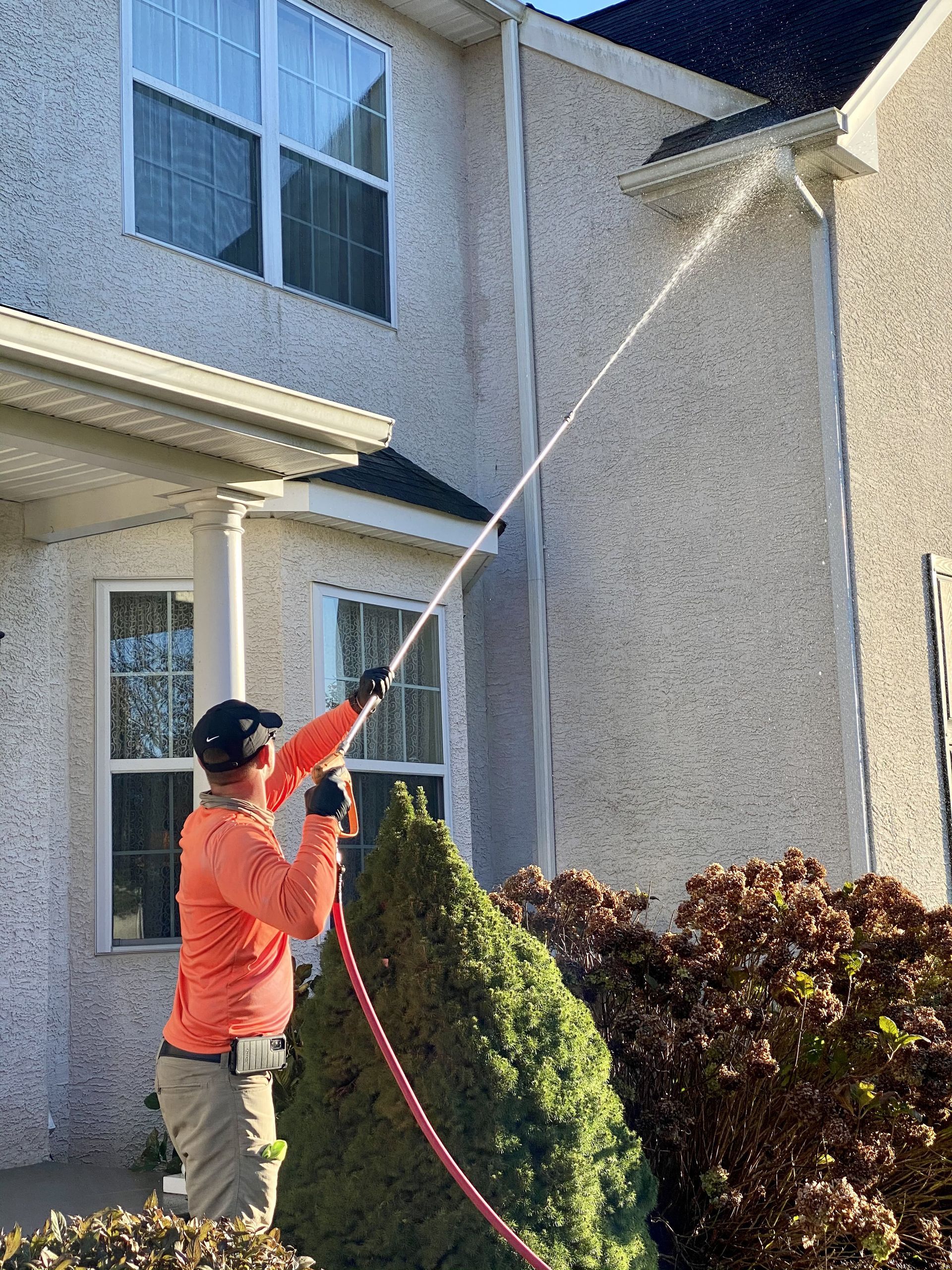 A worker in an orange long-sleeved shirt uses a power washer to spray the exterior of a light-colored stucco home.