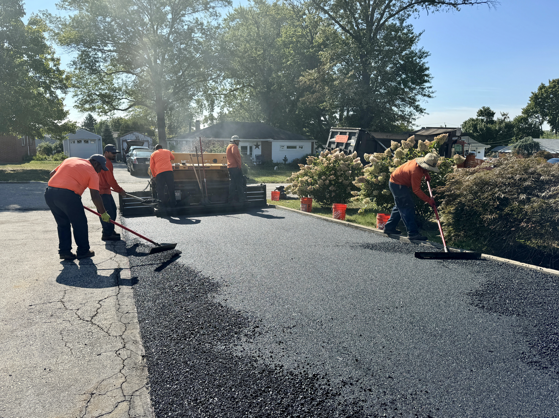 Workers in orange safety shirts pave a residential driveway with fresh asphalt under a sunny sky.