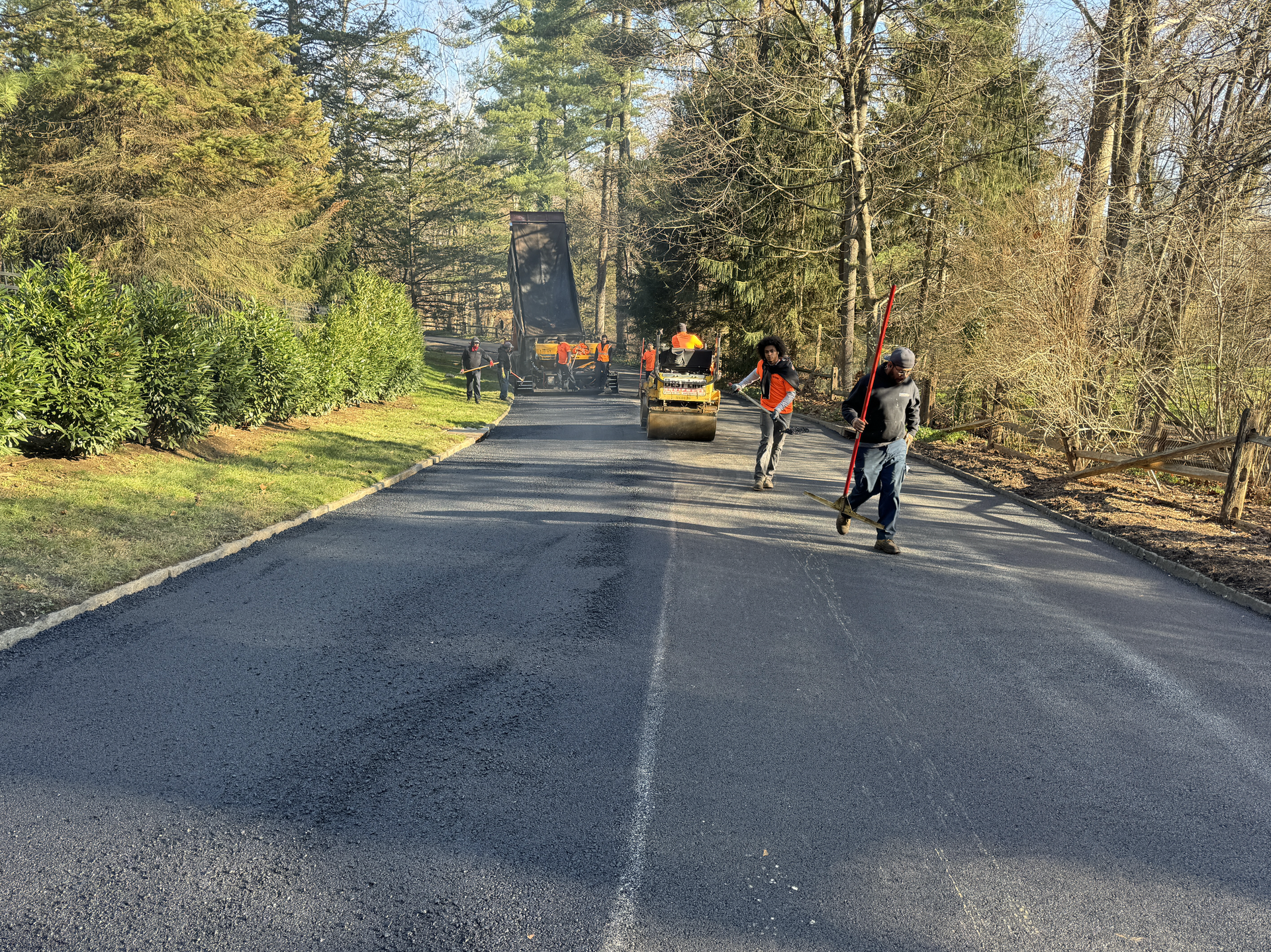 Workers pave a residential driveway with asphalt, using a small roller machine on a sunny day surrounded by trees.
