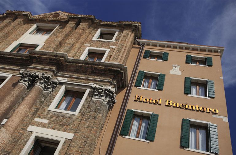 A low-angle view of the tan, multi-story Hotel Bucintoro building attached to a stone facade with Corinthian columns.
