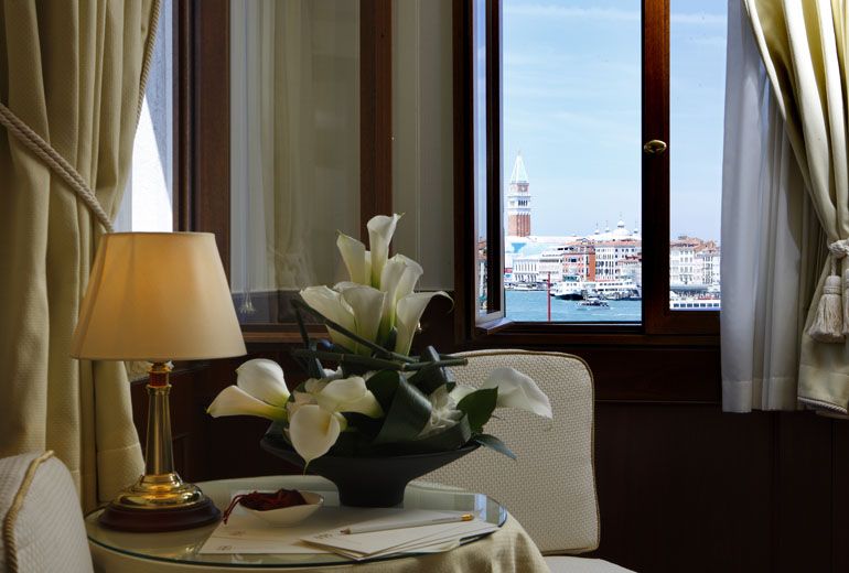 A table with a lamp and calla lily bouquet sits by a window overlooking a Venetian canal and the San Marco campanile.