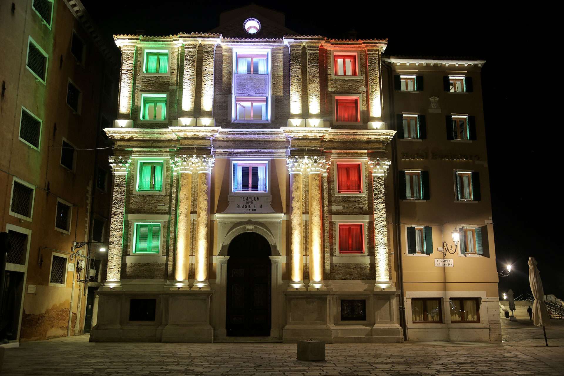 A historical building facade illuminated with green, white, and red lights in a square at night in Venice, Italy.