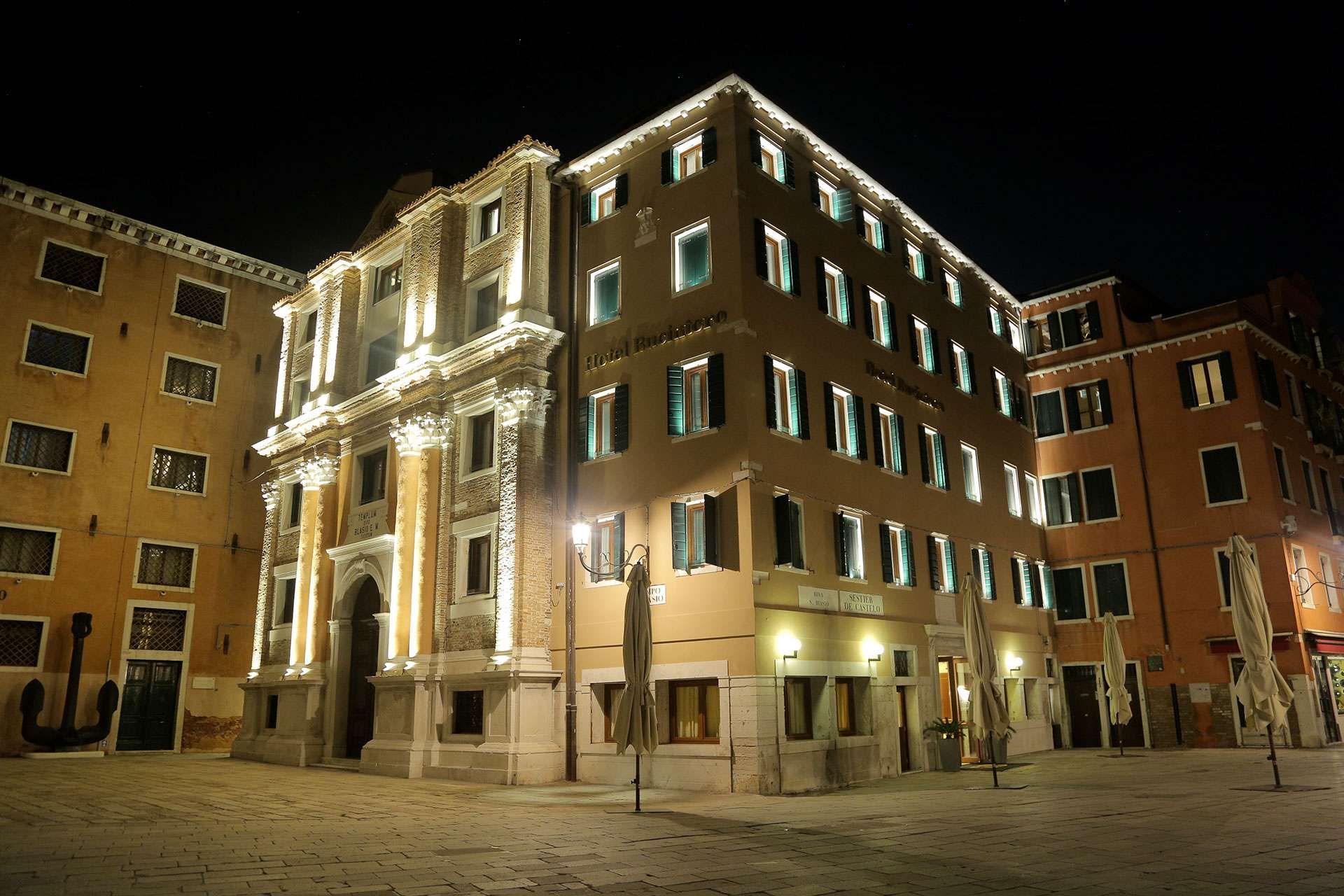 The illuminated facade of a tall, beige historic hotel in a Venetian piazza at night, with dark shuttered windows.