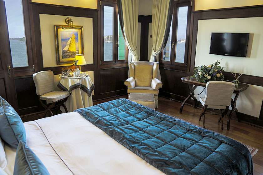 A luxury hotel room with a large bed, seating area, desk, and windows offering a view of the water.